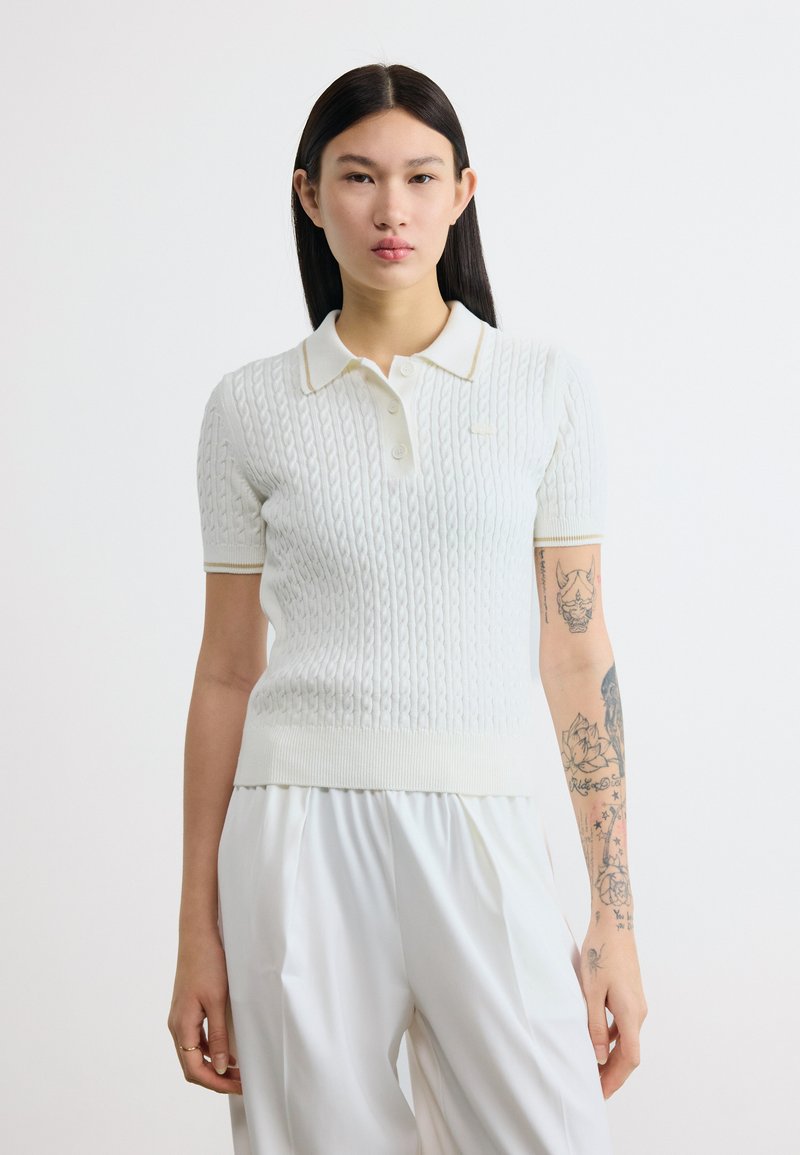 Νεαρή γυναίκα με μακριά σκούρα μαλλιά, φορώντας λευκό πόλο με κοντά μανίκια και πλεχτό σχέδιο, και λευκό παντελόνι, που δείχνει το τατουαρισμένο δεξί της χέρι.