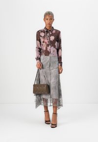Gjennomsiktig brun blomstermønstret bluse, grå blonde midi-skjørt, holder en teksturert brun håndveske, og har på seg svarte sandaler med stropper.
