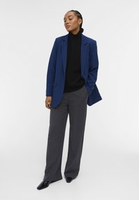 Marineblauwe blazer, zwarte coltrui en grijze wijde pantalon. Gladde stof met een getailleerde pasvorm en scherpe revers. Glanzende zwarte schoenen.