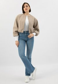 Béžová zkrácená bomber bunda s balónovými rukávy, se zipem; nošena přes bílý top, v kombinaci se světle modrými zvonovými džíny a bílými teniskami.