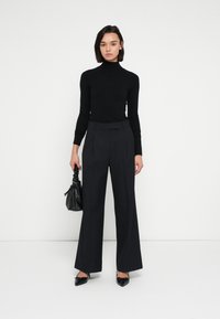 Čierny rolák spojený s nohavicami s vysokým pásom a čiernym prúžkom. Modelka drží malú čiernu kabelku a má na sebe čierne špicaté topánky.