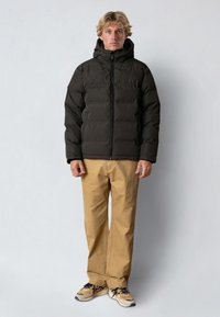 Giubbotto imbottito nero con cappuccio, chiusura con zip e texture a righe orizzontali, abbinato a pantaloni color cammello e sneakers multicolori.