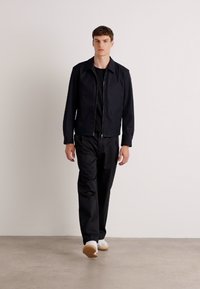 Casaco preto com fecho éclair combinado com calças pretas; modelo usando sapatilhas brancas; tecido suave; design minimalista sem padrões visíveis.