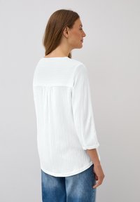 White textured blouse with a rounded neckline and three-quarter sleeves. Features a yoke back and relaxed fit, paired with blue denim.