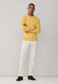 Geel kabelgebreid sweater, witte broek, donkerbruine schoenen. De sweater heeft een gestructureerd patroon, geribbelde mouwen en kraag.