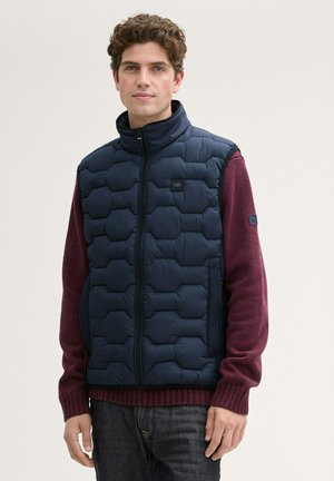 Navy quilted puffer vest with hexagonal pattern, high collar, front zipper, side pockets, worn over a burgundy sweater, paired with dark denim jeans.