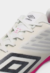 Λευκό αθλητικό παπούτσι Umbro με μαύρο λογότυπο, λευκούς κορδόνια, υφασμάτινο πάνω μέρος και ροζ λεπτομέρειες κοντά στη σόλα.