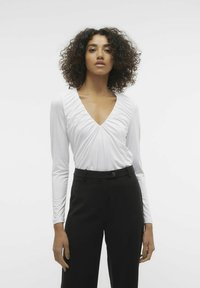 Blusa bianca a maniche lunghe con scollatura a V profonda, caratterizzata da dettagli arricciati nel colletto. Abbinata a pantaloni neri a vita alta.