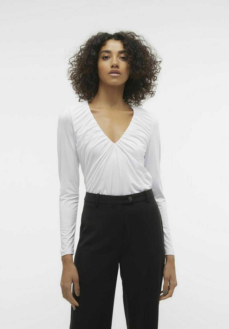 Blusa bianca a maniche lunghe con scollatura a V profonda, caratterizzata da dettagli arricciati nel colletto. Abbinata a pantaloni neri a vita alta.