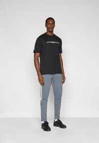 Μαύρο κοντομάνικο t-shirt με το λευκό γράμμα "JUVENTUS" τυπωμένο, σε συνδυασμό με γκρι παντελόνια φόρμας με λευκές ρίγες στο πλάι και μαύρα αθλητικά παπούτσια.