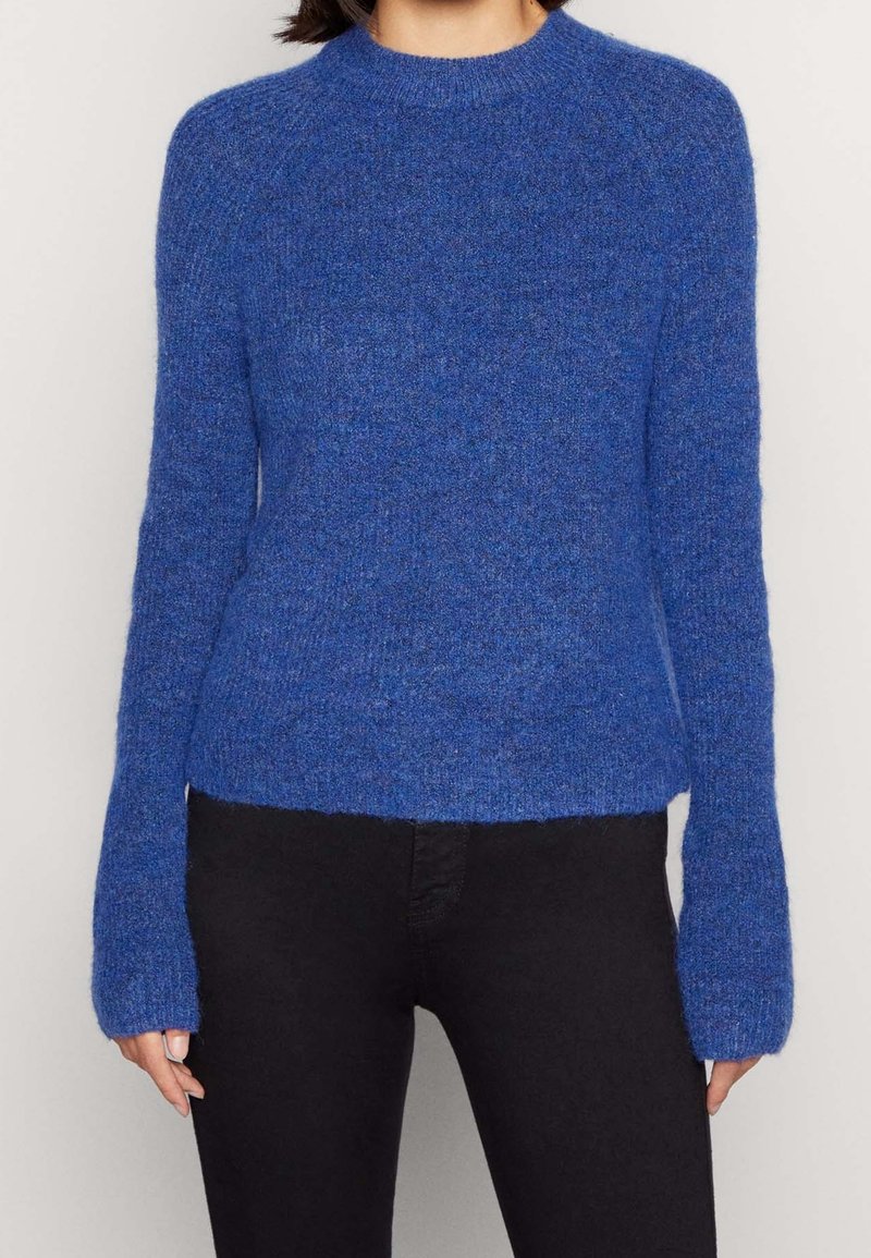 Personne portant un pull tricoté bleu à manches longues associé à un pantalon noir, montré des épaules aux hanches sur un fond uni.