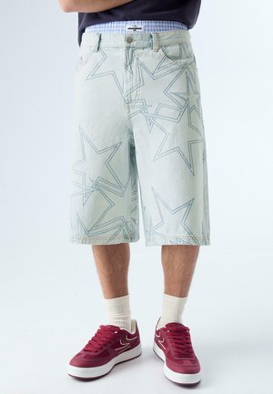 Férfi világos színű farmer rövidnadrágban, csillagok körvonalával, fehér bordázott zokniban, valamint bordó sportcipőben, fehér talppal.