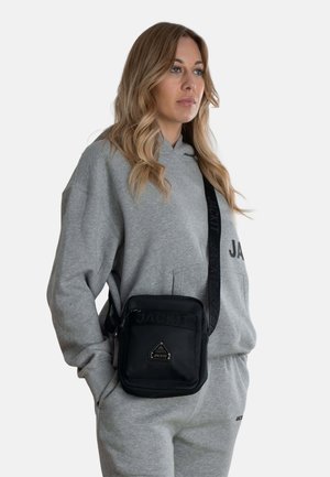 Borsa a tracolla nera in tessuto con logo impressa, dotata di chiusura con zip e tracolla regolabile. Indossata sopra una felpa grigia e pantaloni.