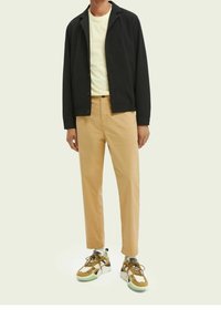 Schwarze Knopfjacke, hellgelbes T-Shirt, beige Hose und mehrfarbige Sneakers mit Akzenten in Grün und Braun. Einfacher, lässiger Look.