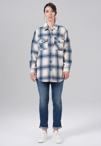 Een geruite flanellen shirt in blauw- en beigetinten, met knoopsluiting en twee borstzakken. Gepaard met donkere jeans en witte sneakers.