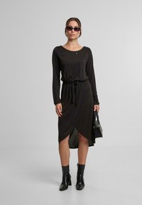 Schwarz langärmliges Kleid mit gebundenem Taillengürtel, Wickeldesign, weicher Textur und mittellanger Länge. Getragen mit schwarzen Stiefeletten und Sonnenbrille.