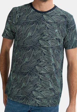Mand iført en mørkeblå t-shirt med et lyst grønt bladmotiv over det hele og mørkeblå jeans, stående mod en ensfarvet baggrund.