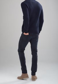 Granatowy prążkowany sweter, dopasowane ciemne jeansy i beżowe zamszowe botki za kostkę. Całość casualowego outfitu widziana od tyłu na neutralnym tle.