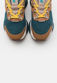 Paire de baskets turquoise et marron avec des lacets jaunes, des œillets en métal, et des semelles épaisses blanches et turquoises sur fond blanc.