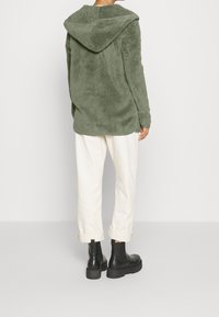 Green fleece jacket with a large hood, featuring a soft texture. Paired with cream-colored cuffed pants and black chunky boots.