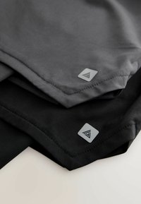 To beklædningsgenstande i gråt og sort stof stablet oven på hinanden. Hver af dem har et elegant design med et sølvfarvet trekantet logo i det nederste hjørne.