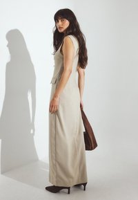 Beige udenærmet kjole med et tætsiddende design, detaljer med sidelommer og en lang, svævende silhuet. Modellen holder en brun håndtaske og står foran en neutral baggrund.