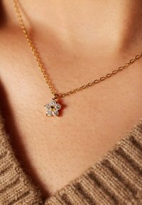 Gouden ketting met een klein bloemenvormig hangertje versierd met zes heldere edelstenen, tegen een huidskleurige achtergrond.