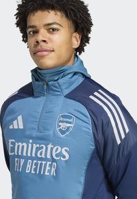 Młody mężczyzna w niebieskiej kurtce piłkarskiej Arsenal z logo Emirates Fly Better oraz paskami Adidas, patrzący do przodu z neutralnym wyrazem twarzy.