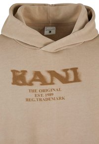 Hoodie beige avec un grand logo "KANI" légèrement effacé en marron. Doté d'une capuche avec cordon de serrage et d'une étiquette visible à l'intérieur de l'encolure.