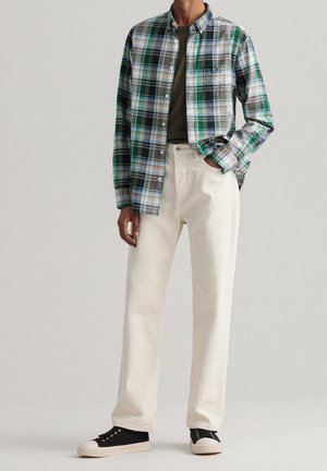 Persona con camisa de cuadros verde y negra sobre camiseta verde oscuro, pantalones crema y zapatillas negras con suelas blancas, de pie con la mano en el bolsillo.