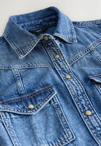 Džínsová košeľa s klasickým golierom, dvoma prednými vreckami, zapínaním na gombíky, modrou látkou a kontrastnými švami.