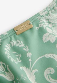 Zakladni material v mint zeleni barvi z belimi cvetličnimi motivi. Ima zlato dekorativno oznako pritrjeno z nebesno modro nitjo.