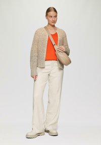 Beige gestrickte Strickjacke über einem orangefarbenen Oberteil, kombiniert mit cremefarbenen, weit geschnittenen Hosen und beigen Slippern. Hält eine kleine beige Umhängetasche.