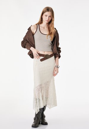 Jeune femme portant un crop top rayé, une veste marron posée sur les épaules, une jupe en dentelle blanche avec une ceinture marron, et des bottes noires, debout devant un fond blanc.