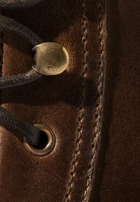Primo piano di un stivale in pelle marrone con lacci neri, occhielli metallici tondi e accessori in ottone. I dettagli delle cuciture visibili esaltano la texture.