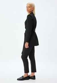 Blazer noir ajusté avec une taille ceinturée, associé à un pantalon slim noir et des mocassins en cuir noir. Texture lisse, design minimaliste.
