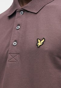 Bordeauxrotes Poloshirt mit strukturierter Stoffqualität, drei Knöpfen und einem gestickten gelben Vogel-Logo auf der linken Brustseite. Verfügt über verstärkte Nähte.