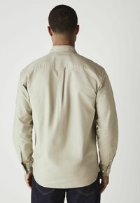 Lichtbeige overhemd met lange mouwen en knoopmanchetten, achterste schoudernaaddetail en rechte zoom. Draag het zoals gezien van achteren, gecombineerd met donkere jeans.