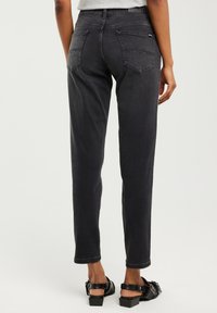 Osoba v černých džínách s vysokým pasem a zadními kapsami a černých sandálech na podpatku na jednobarevném světlém pozadí.