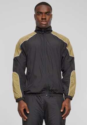 Black zip-up jacket with olive green shoulder panels and white piping. Made of lightweight material, featuring elastic cuffs and a fitted waist.