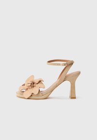 Beige high-heeled sandaal van suede, met een grote bloemenaccent aan de voorkant, verstelbare enkelband en een gesloten neus ontwerp.
