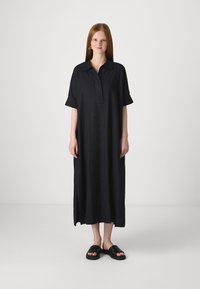 Vestido preto com botões, feito de tecido leve, apresentando um decote com colarinho, mangas curtas e fendas laterais. Usado com sandálias pretas simples.