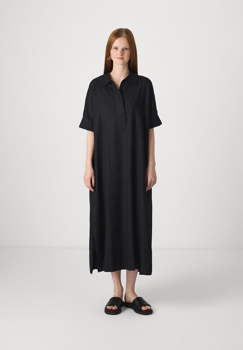 Vestido preto com botões, feito de tecido leve, apresentando um decote com colarinho, mangas curtas e fendas laterais. Usado com sandálias pretas simples.