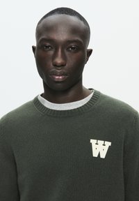 Olive green sweater with ribbed collar and cuffs, featuring a white “W” logo on the chest. Made from textured knit material.