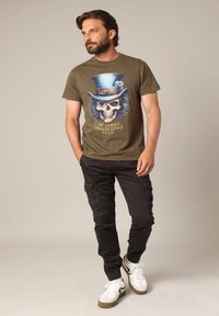 T-shirt vert olive avec un graphique de crâne et de haut-de-forme, associé à un pantalon cargo noir et des baskets blanches avec des accents sombres.
