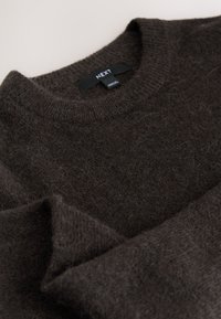 Donkerbruine trui gemaakt van zacht, pluizig materiaal. Heeft een ronde halslijn en een geribbelde kraag. Merkentag zichtbaar in het midden.