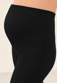 Leggings neri a coste con vita elastica ampia. La texture è liscia con linee verticali, evidenziando una silhouette aderente.