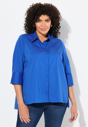 Blouse bleu à col avec une fermeture à boutons, des manches trois-quarts et une texture lisse. Portée avec un jean en denim foncé sur un fond uni.
