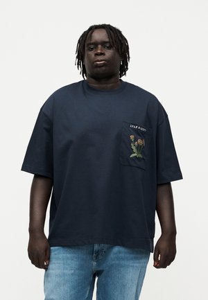 Mand med dreadlocks iført en navy oversized T-shirt med blomstermotiv og teksten "LYLE & SCOTT" på brystlommen, kombineret med lyseblå jeans.