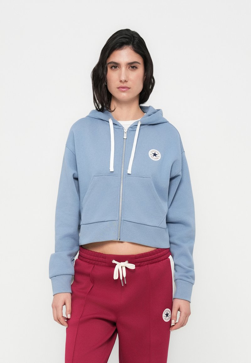 Sweat à capuche court bleu clair avec fermeture éclair, poche avant et cordons blancs, associé à un pantalon de jogging bordeaux avec un patch logo.